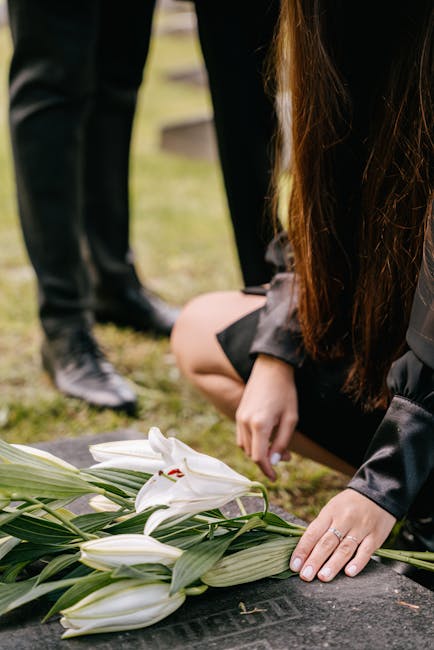 A person dressed in black, kneeling on the ground, arranging a bouquet of white lilies with large, lush green leaves, in an outdoor setting with soft natural lighting. The lilies have smooth, curved p