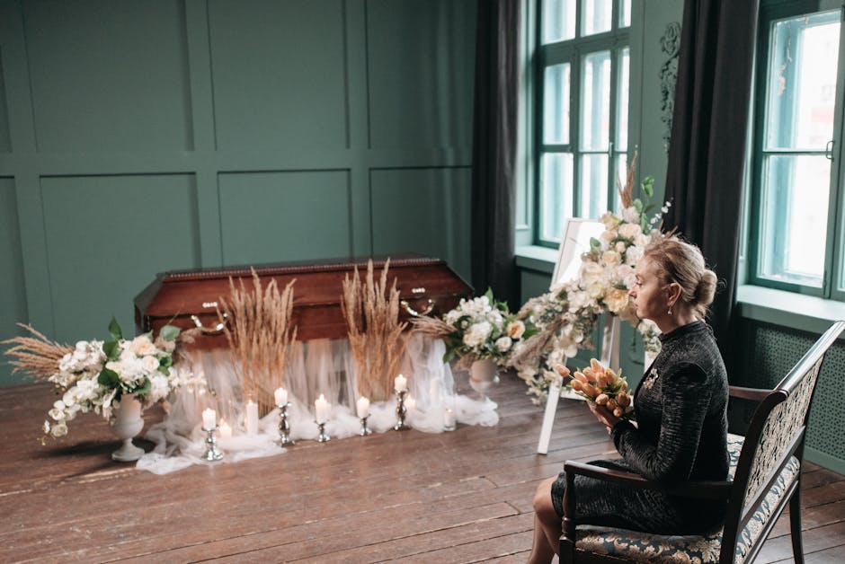 A woman seated on a vintage armchair in a softly lit room, holding a bouquet of orange and cream roses. In front of her, there is a funeral floral arrangement on a wooden table, featuring white roses,