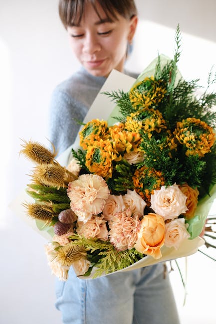 A woman is holding a large, vibrant floral arrangement featuring a mix of fresh flowers including peach roses, yellow and orange marigolds, and pink carnations, complemented by green foliage and textu