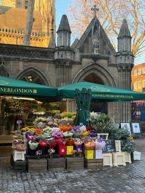 A vibrant flower stall situated in front of a historic stone building with Gothic architectural features, including pointed arches and decorative pinnacles. The stall, protected by a green canopy mark