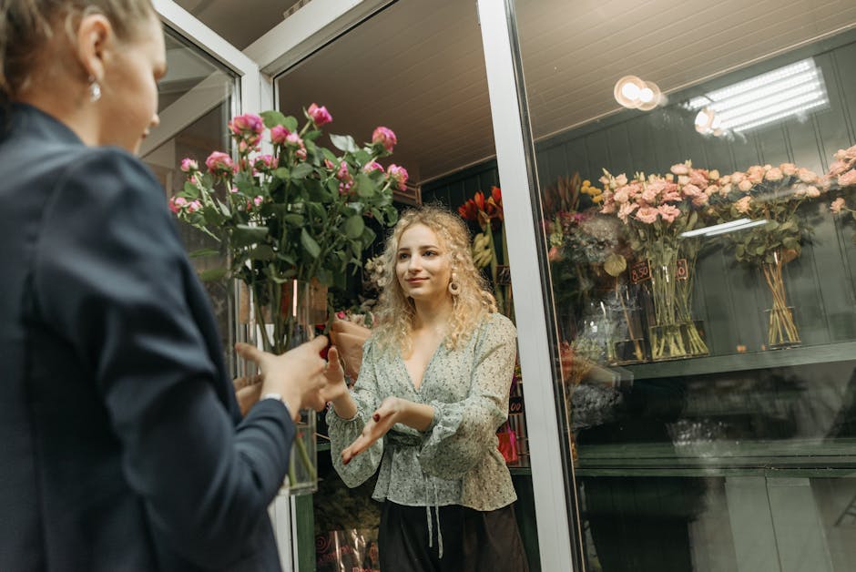 A woman inside a flower shop with a glass display case filled with pastel pink and peach roses arranged in tall, clear glass vases. She has curly blonde hair, wears a floral-patterned light green blou