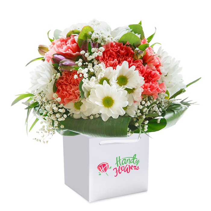 A strikingly arranged bouquet, radiating both warmth and cheer, is gracefully presented upright within a crisp white square gift bag-ideal for easy transport or delivery throughout South Croydon. Rich coral and peach carnations form the heart of this floral composition, their abundant, frilled petals lending a luxurious softness. Bright white daisies, each with a vibrant lime-green center, nestle between the larger blooms, bringing joyful contrast reminiscent of the lively atmosphere along Brighton Road. Delicate clouds of baby's breath add an ethereal, airy quality, while several closed, purple-tipped buds-perhaps early spring alstroemeria-punctuate the design, hinting at the unfolding beauty of local botanical gardens. The entire bouquet is encircled by lush, deep green leaves, their glossy surfaces providing structure and serenity, much like the leafy lanes of nearby Purley Oaks. The sturdy white gift bag, complete with elegant ribbon handles, is as inviting as it is practical, making this arrangement perfect for delivery to loved ones across South Croydon or as a centrepiece for a special celebration at home. Photographed on a pure white background, the colours and textures of each bloom are brought into sharp, vibrant focus, embodying a sense of freshness and abundance that reflects the expertise of local florists.