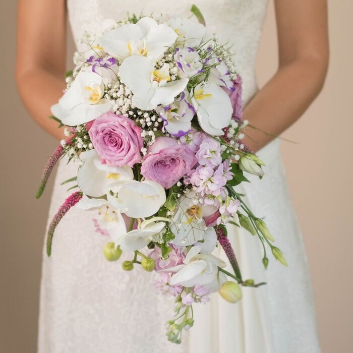 This exquisite bridal bouquet, crafted with passion near South Croydon's bustling High Street, radiates a sense of serene poise and gentle celebration. The bride, visible from mid-torso, is enveloped in a gown of ivory silk and intricate lace-a fitting canvas for the opulent, waterfall-style bouquet that dominates the foreground. At the centre, clusters of full, lavender and blush pink roses open majestically, bordered by the striking presence of white Phalaenopsis orchids whose broad petals and subtle golden centres catch the soft ambient light. Wispy baby's breath and tiny lavender-edged blooms interweave with textured amaranthus, its fuchsia tendrils tumbling downward in plush, tactile trails, adding movement and drama. Subtle greenery and emerging buds peek out between the larger flowers, recalling strolls through the shaded paths of Haling Grove. The interplay of forms, from cloud-like fillers to the structured drama of the orchids, achieves a sense of both classic romanticism and modern artistry. The colour palette-pristine whites, powdery pinks, serene lavenders, with the faintest hint of burgundy from trailing accents-captures the mood of a spring or summer morning wedding in the local community. Every element, from the bouquet's cascading silhouette to its gentle fragrance, speaks to the expert skills of local florists in South Croydon. This bouquet doesn't simply complement a bride-it embodies a love story unfolding in every petal.