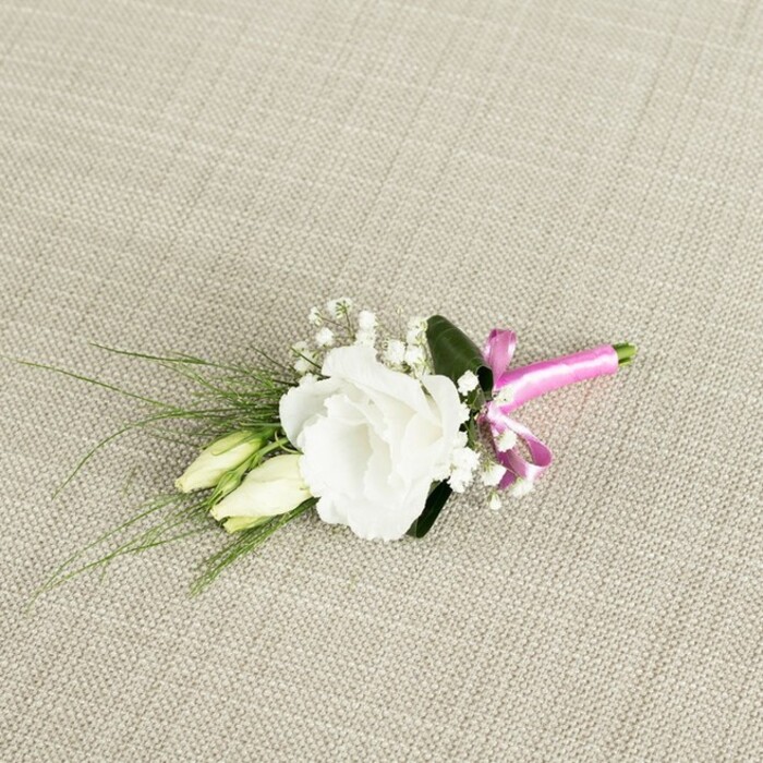 A thoughtfully composed boutonnière is shown resting at a gentle diagonal on a warm, light beige fabric with a distinct woven texture, reminiscent of fine linen or soft burlap laid out on a dressing table before a ceremony in South Croydon. At the centre of the arrangement, a single white eustoma flower is fully open, its many ruffled petals curving and folding in soft, irregular layers that give it a luxurious, almost rose-like fullness. The bloom sits just off-centre to the right, drawing the eye immediately with its clean, bright tone and subtle shadows between the petals. Nestled close beside it, slightly below and to the left, are two elongated lisianthus buds, still tightly closed and shaded in creamy white and pale green, suggesting flowers that will open later in the day as the celebration continues. Fine, cloud-like clusters of white baby's breath are scattered throughout the small composition, their tiny, bead-sized flowers adding volume and a hint of romance without overwhelming the central bloom. From behind, slender, grass-like blades of greenery splay out towards the left, some curving gently, creating a sense of movement and echoing the relaxed greenery found in local gardens and green spaces near Purley Oaks. A single, broader dark green leaf is tucked just behind the eustoma, providing a backdrop that intensifies the contrast with the white petals and helps anchor the design visually. At the base, the stems are neatly gathered, then wrapped with a smooth, fuchsia satin ribbon tied into a compact, playful bow, the vivid pink adding a contemporary, celebratory twist to the classic white and green palette. The lighting is soft and even, revealing every texture-from the fabric's weave to the satin's sheen-making this boutonnière appear fresh, handcrafted, and ready to be pinned to a lapel for a wedding, prom, or formal event in and around South Croydon.
