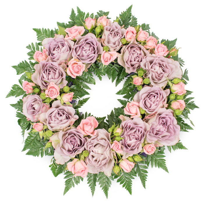 Viewed from directly overhead, a lush condolence wreath fills the frame, its circular outline sharply defined against a smooth white backdrop. The arrangement is composed with careful symmetry, yet it feels organic and flowing, as though the roses have naturally grown into this ring of remembrance. Dominating the design are large, open roses in an elegant dusty lavender and muted mauve tone, each bloom with layers of softly curling petals that form deep, cup-like centres. The colour is understated, almost vintage in character, giving the wreath a timeless, refined presence. Nestled between these larger roses are small clusters of blush pink spray roses and rosebuds, their lighter, fresher colour bringing a gentle warmth to the cooler purples. Some of these miniature roses are fully open, revealing tight clusters of tiny petals, while others are still part-closed, suggesting new memories held alongside older, cherished ones. Beneath and around the blooms, a thick base of dark green, finely divided fern fronds creates a strong, textural foundation, the serrated leaves radiating outward in overlapping layers. Occasional touches of deeper filler foliage and tiny dark buds are visible in the gaps, subtly deepening the overall colour palette and drawing the eye inward. The light is soft and diffused, enhancing the velvety surfaces of the petals and the satin sheen on the greenery without creating harsh contrast. Designed in the style we often use for South Croydon families saying goodbye in areas like Warlingham, Kenley, or along Addington Road, this wreath conveys grace, respect, and quiet condolence, suitable for a funeral service, memorial gathering, or celebration of life.