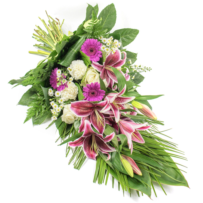 This photograph shows a beautifully crafted funeral sheaf laid against a clean white background, its diagonal, crescent-like form creating a sense of gentle movement from left to right. At the upper left, a cluster of neatly bound stems marks the starting point, then the flowers sweep downwards into a lush cascade of colour and texture. Several prominent Stargazer lilies dominate the centre, their petals a vivid fuchsia-pink with crisp white edges and dark speckling, opening outward as if in a final, gracious gesture of farewell. Around these lilies, bright magenta gerbera daisies form cheerful yet poignant circles of colour, suggesting treasured memories and affection. Soft white roses, in stages from tight bud to partially open, are interwoven with the lilies, bringing an atmosphere of purity, grace and enduring love. Between them, white carnations with ruffled petals add volume and a velvety softness, while tiny white daisy-like flowers peek through the larger blooms, adding small points of light throughout the design. The arrangement is richly supported by deep green foliage: broad leaves act almost like a cradle, holding the flowers together, while airy fern fronds spill outward along the sides. Long, slender leaves extend towards the bottom right, emphasising the elongated shape and guiding the viewer's gaze along the whole tribute. The composition has the careful balance and gentle structure of a piece created by an experienced florist at Flowers South Croydon, accustomed to preparing respectful tributes for local services in areas like Purley Way, Lloyd Park, and Sanderstead. It conveys a mood of solemn beauty and quiet consolation, ideal for placing beside a casket, on a memorial table, or at a graveside as a tender expression of sympathy.