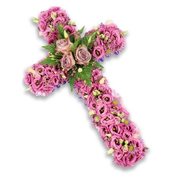 This image shows an intricately designed floral cross tribute placed against a stark, pure white background, the soft drop shadow giving the impression that the cross is gently lifted from the surface. The structure is completely covered with fresh flowers, creating a lush, uninterrupted tapestry of soft pinks, mauves, and muted purples. At the centre, where the vertical and horizontal bars intersect, three larger roses in a sophisticated dusty rose or light mauve-pink shade form the focal point, their open, cupped blooms displaying layers of velvety petals. Encircling these roses are fine, arching fronds of bright green fern and smaller, deeper green leaves, which add contrast and make the central cluster stand out. Extending along the arms and down the length of the cross, numerous lisianthus (Eustoma) flowers are tightly packed together, their ruffled, trumpet-shaped petals ranging from pale blush to more vivid pink, with some blooms revealing darker, almost berry-toned centres. Among them, smaller filler flowers in pale pink, lilac, and soft white introduce delicate texture, while tiny yellow or cream centres catch the light. Here and there, light green buds peek through, hinting at blossoms yet to open and adding a gentle sense of growth amid remembrance. Subtle glimpses of a darker purple or blue base, likely ribbon or structural material, are visible at the very edges, lending definition to the cross's outline. The overall effect is one of refined beauty and solemn grace, the kind of meaningful tribute that might be chosen by South Croydon families for a funeral or church service along Brighton Road or near local resting places, offering a visual expression of faith, love, and lasting memory.