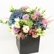 A lavish, modern bouquet from a South Croydon florist shines in a minimalist matte black square container, dramatically placed against a white background to highlight every vibrant detail. Grand clusters of deep indigo and periwinkle hydrangeas dominate, their plush, abundant petals reminiscent of the blooming borders found in local parks. Nestled among them are velvety lavender and mauve roses, each bud in various stages of bloom, expressing gentle romance and layered beauty. Creamy white lisianthus add a soft, classic elegance, their unfurled petals and slender buds reaching gracefully upward. Bright lime green trick dianthus offer an unexpectedly lively texture, their curious shapes popping amidst soft pastels, while slender sprays of feathery pale pink astilbe lend airy movement reminiscent of a gentle Croydon breeze. Glossy leafy greens, varying from fresh lime to deep woodland, weave throughout the composition, enhancing its fullness and evoking the vitality of South Croydon gardens. The arrangement is meticulously designed for all-around enjoyment-its colour palette of cool blues, gentle pinks, creamy whites, and lush greens blending in harmonious balance. Soft, even lighting accentuates every petal, allowing this premium display to radiate warmth and sophistication. Ideal for brightening a home, celebrating a milestone, or delivering heartfelt sympathy throughout South Croydon, this bouquet reflects the discerning taste and passion of local floral designers.